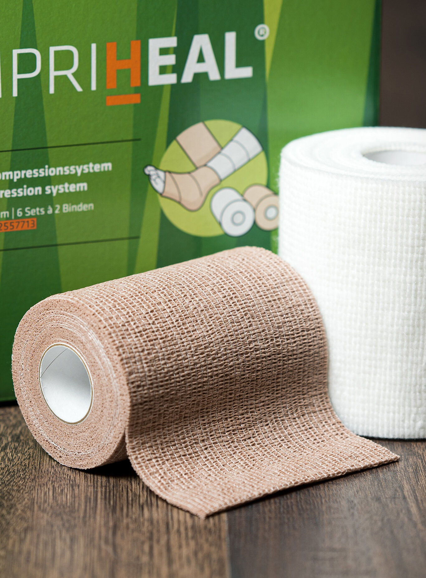 Die Verpackung von Compri-Heal® stehend mit einem Set Kompressionsbinden im Vordergrund auf einem dunklen Holzboden. | © Bamboo Health Care GmbH