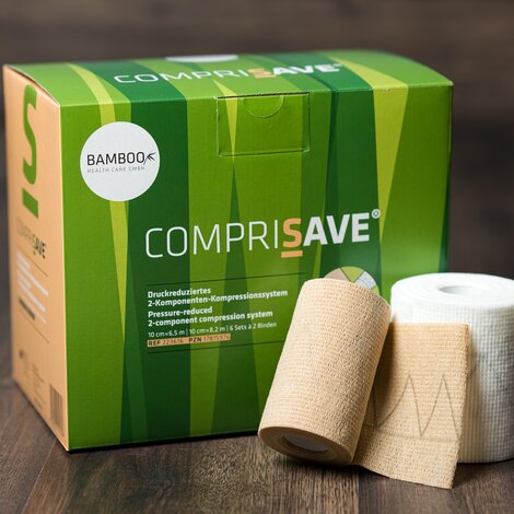 Die Verpackung von CompriSave® stehend mit einem Set Kompressionsbinden im Vordergrund auf einem dunklen Holzboden. | © Bamboo Health Care GmbH