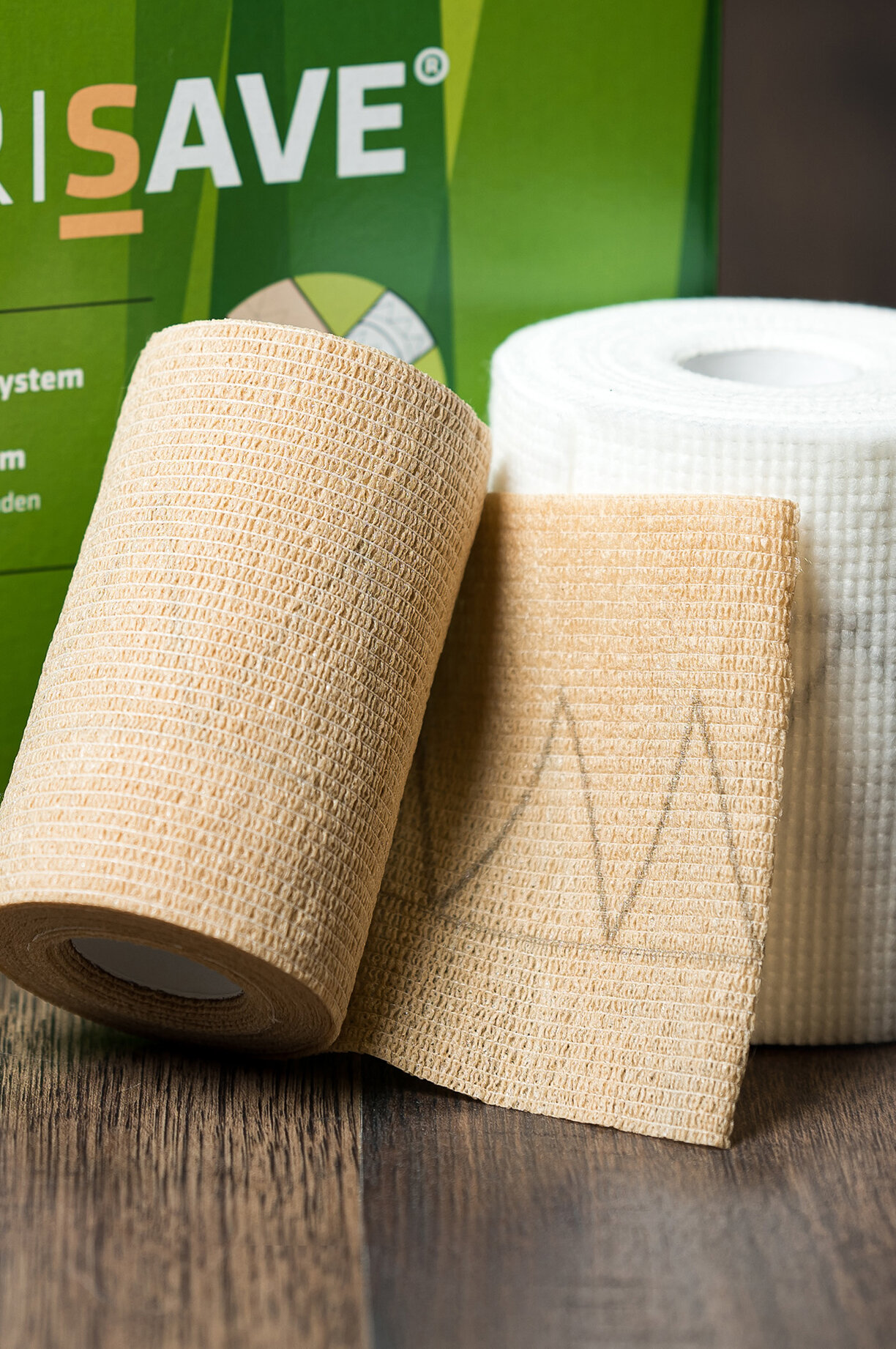 Die Verpackung von CompriSave® stehend mit einem Set Kompressionsbinden im Vordergrund auf einem dunklen Holzboden. | © Bamboo Health Care GmbH