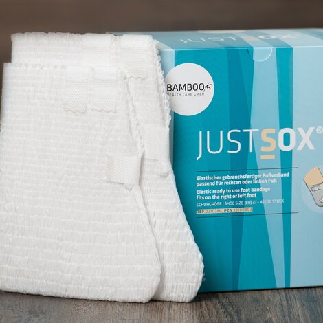 Zwei unterschiedliche Verpackungsgrößen von JustSox® stehend mit zwei Fußverbänden im Vordergrund auf einem dunklen Holzboden. | © Bamboo Health Care GmbH